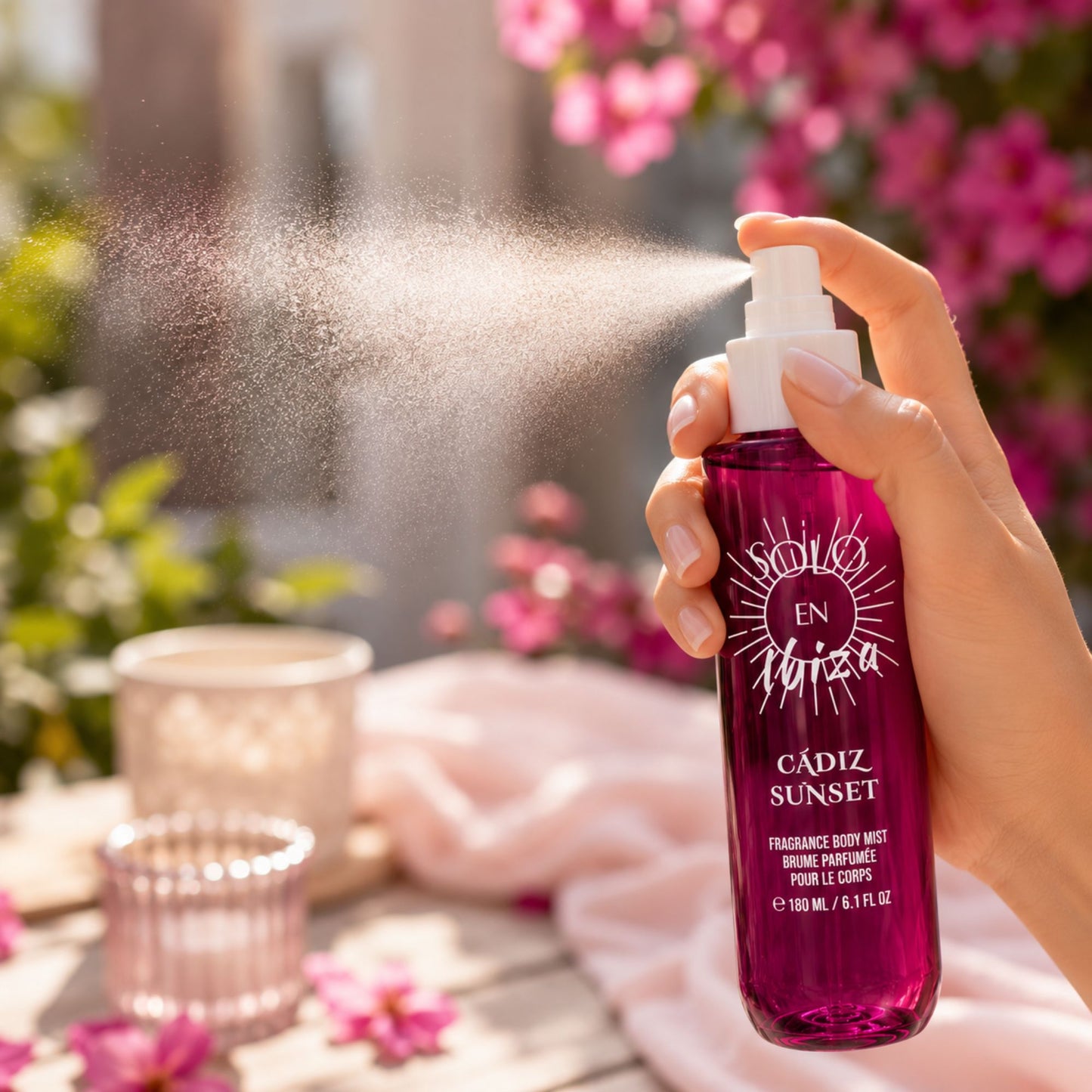 A hand holds a bottle of En Ibiza Cádiz Sunset Fragrance Body Mist, spraying a fine mist in an outdoor garden setting with pink flowers and a soft pink towel in the background.