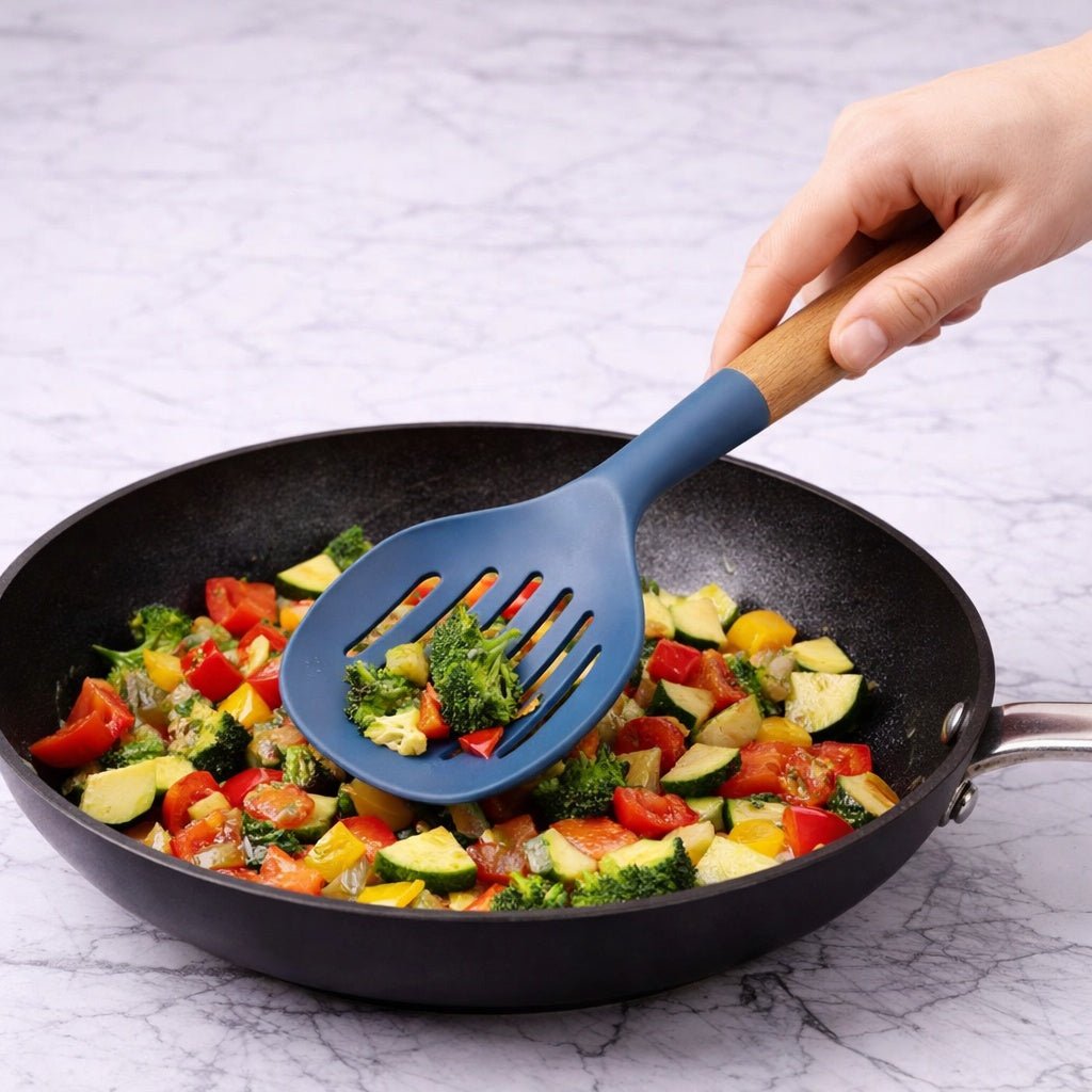 Blue slotted spatula lifting vegetables from a frying pan on a marble surface