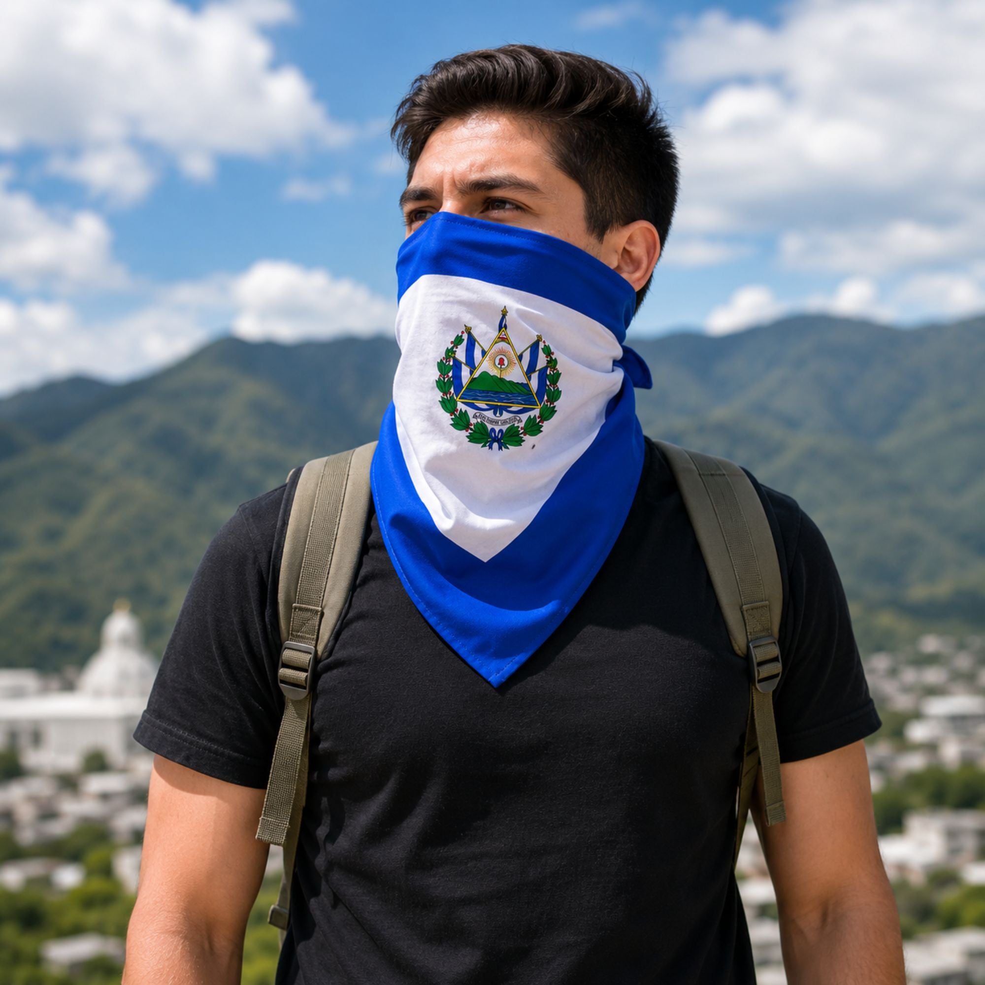 A man wearing a blue and white bandana face covering with the emblem of El Salvador, set against a background of mountains and a blue sky.