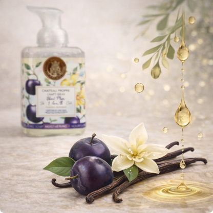 Foaming hand soap bottle with plums, vanilla beans, and a flower on a textured surface.