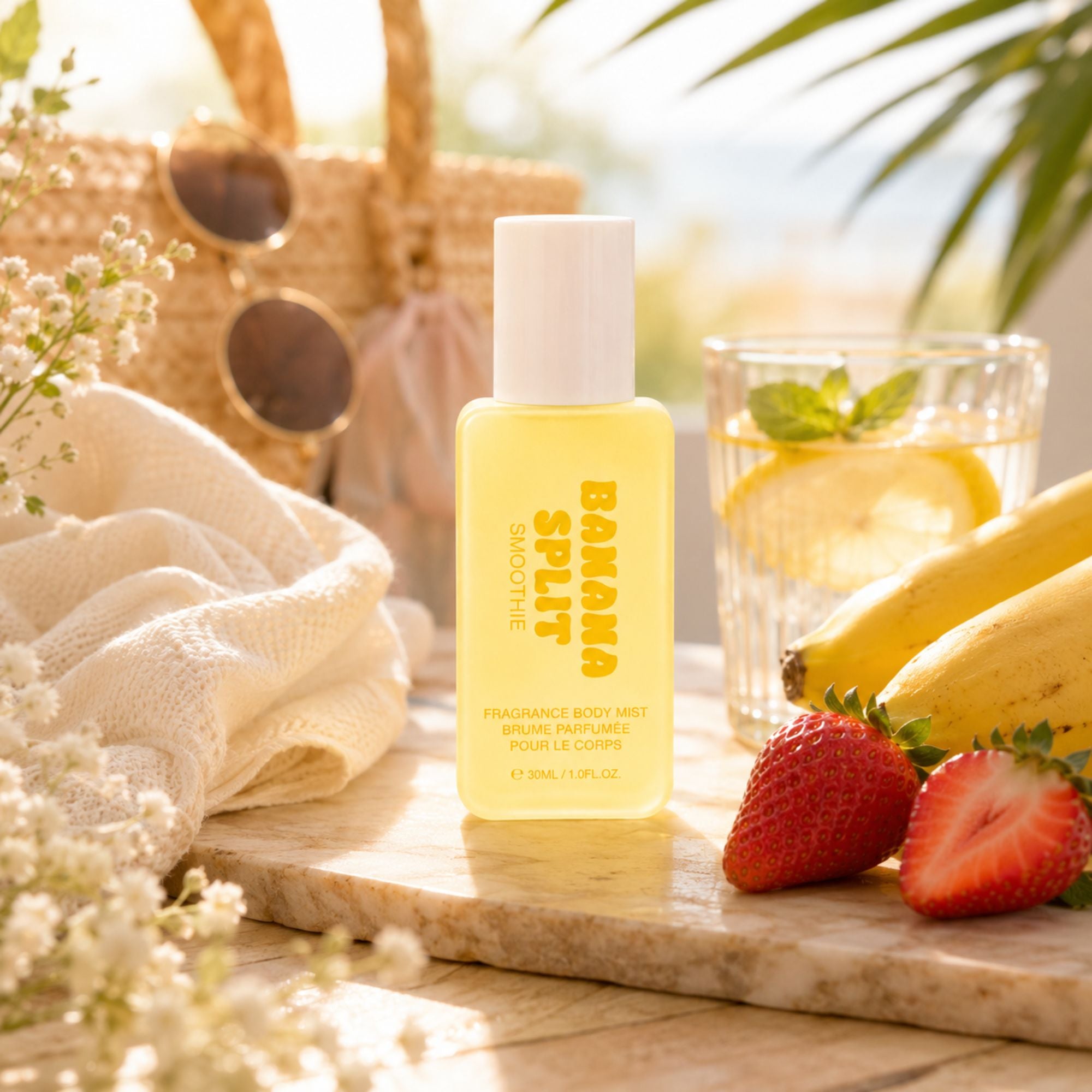 A bottle of Banana Split Fringe Body Mist stands on a wooden board outdoors, surrounded by fresh strawberries, bananas, a glass of lemonade, a woven bag, and a white knit blanket in a warm, sunny setting.