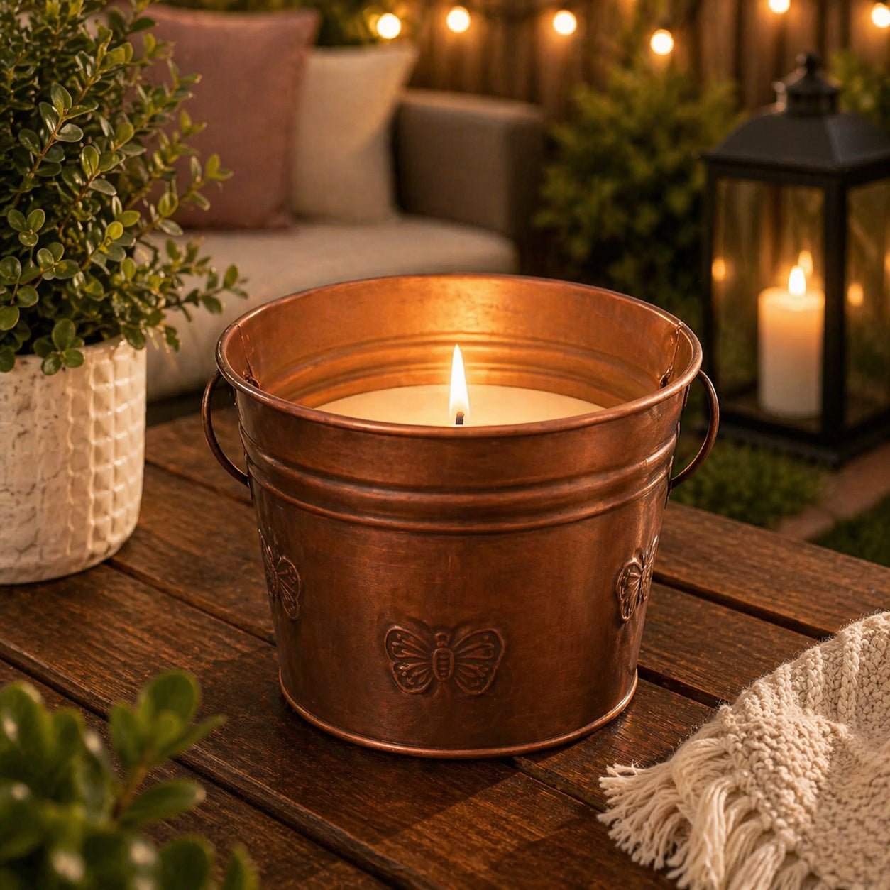 Copper bucket with a lit candle on a wooden surface outdoors, surrounded by plants and a lantern.