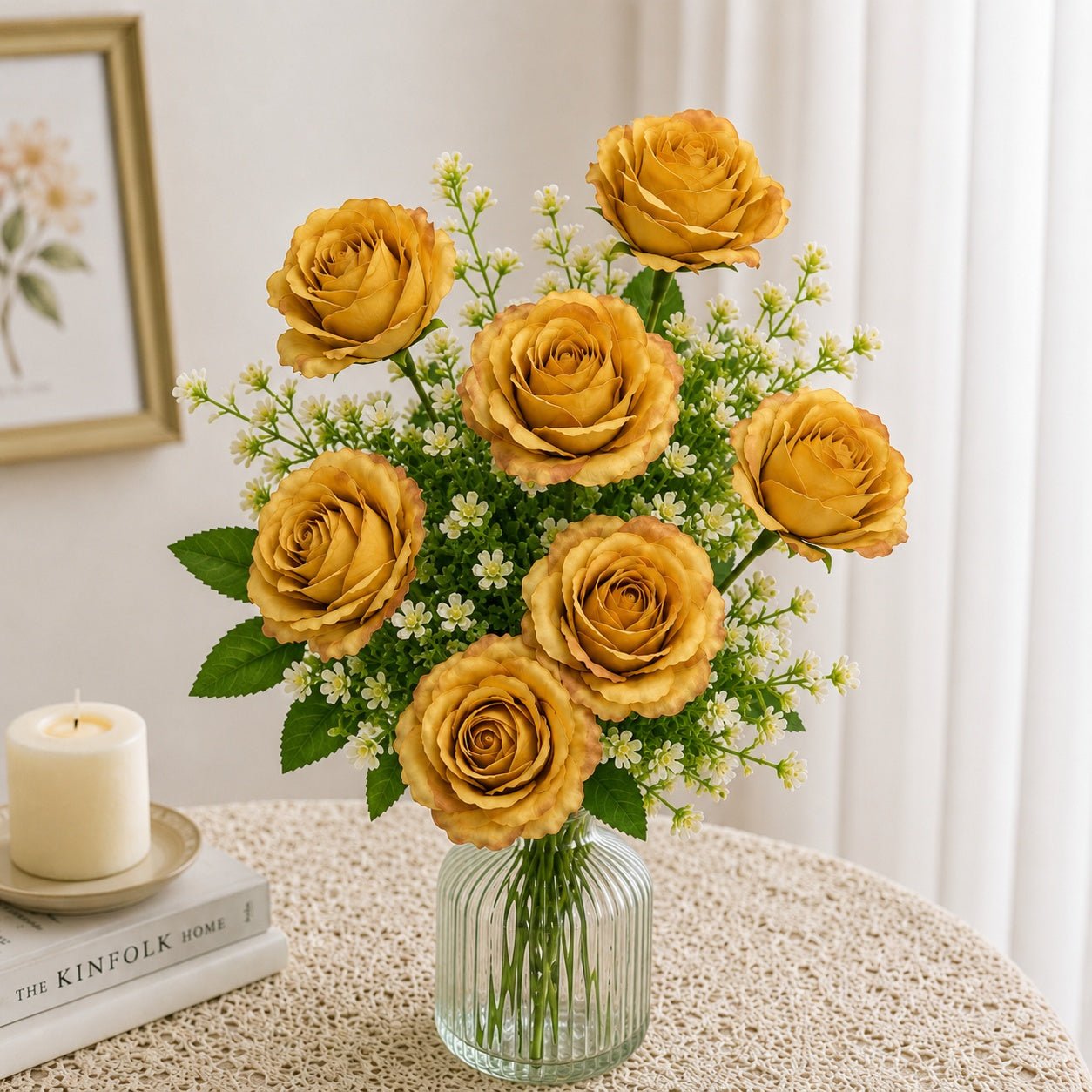 Bouquet of yellow roses in a clear vase on a table with a candle and books in the background.