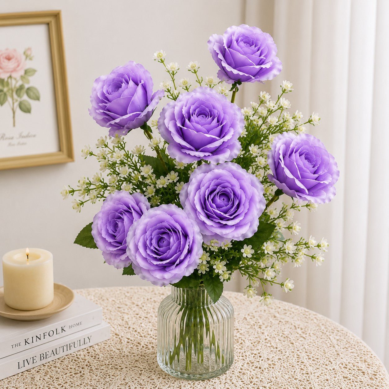 Bouquet of purple roses in a clear vase on a table with a candle and books in the background.
