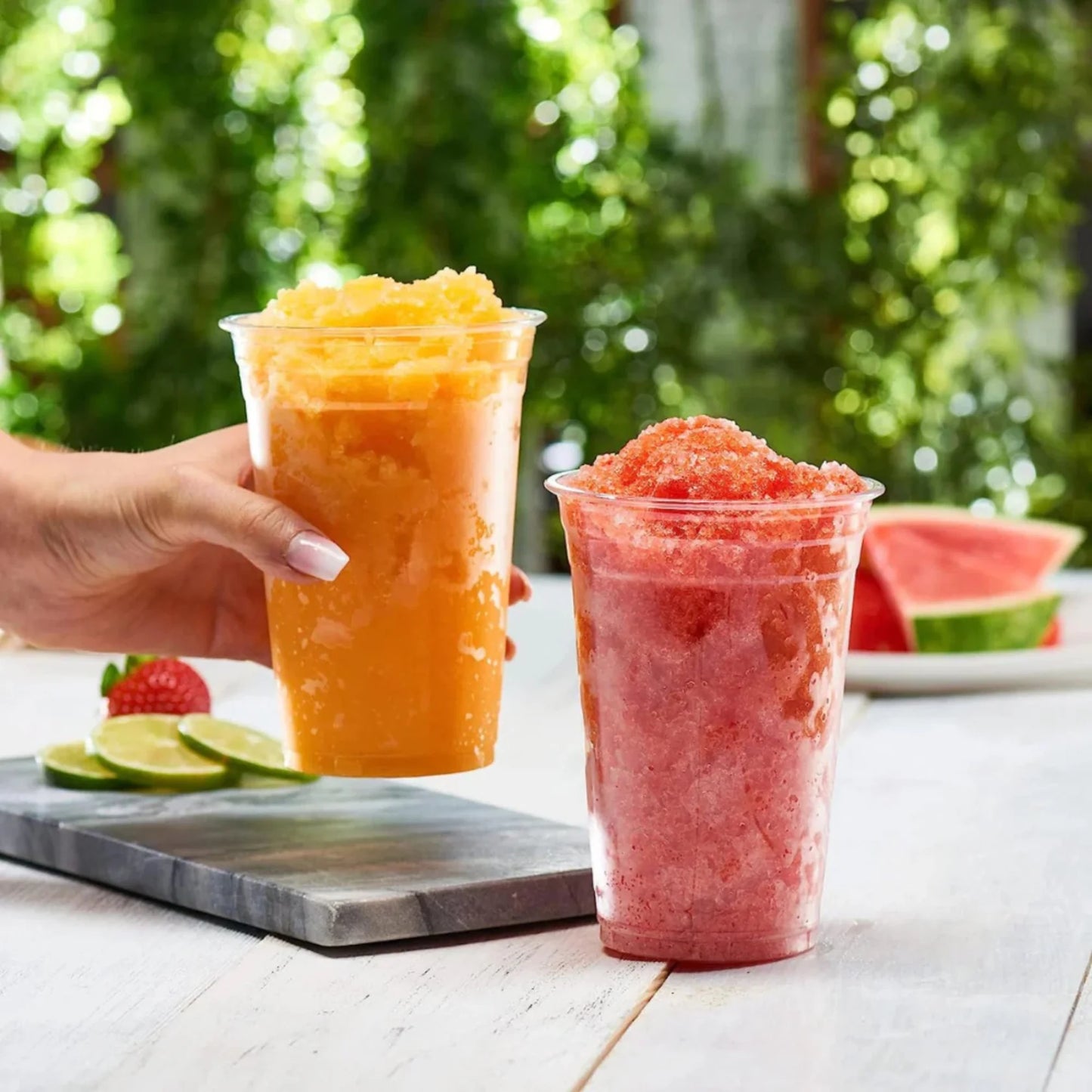 Two cups of colorful slushies on a table with a blurred green outdoor background