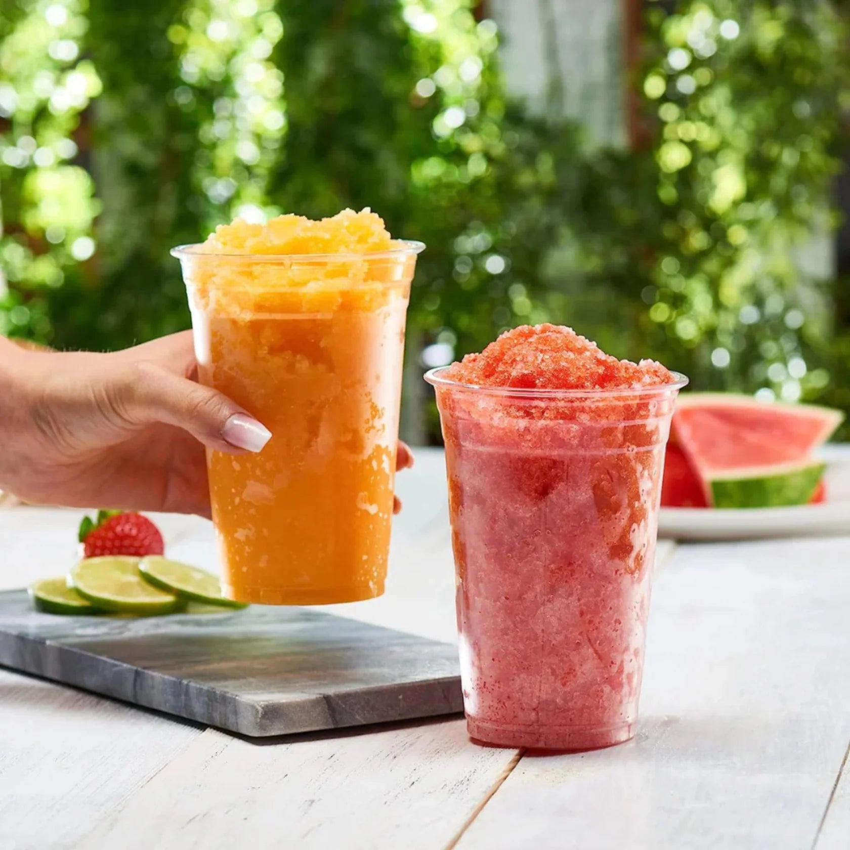 Two cups of colorful slushies on a table with a blurred green outdoor background