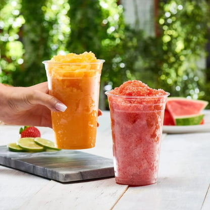Two cups of colorful slushies on a table with a blurred green outdoor background