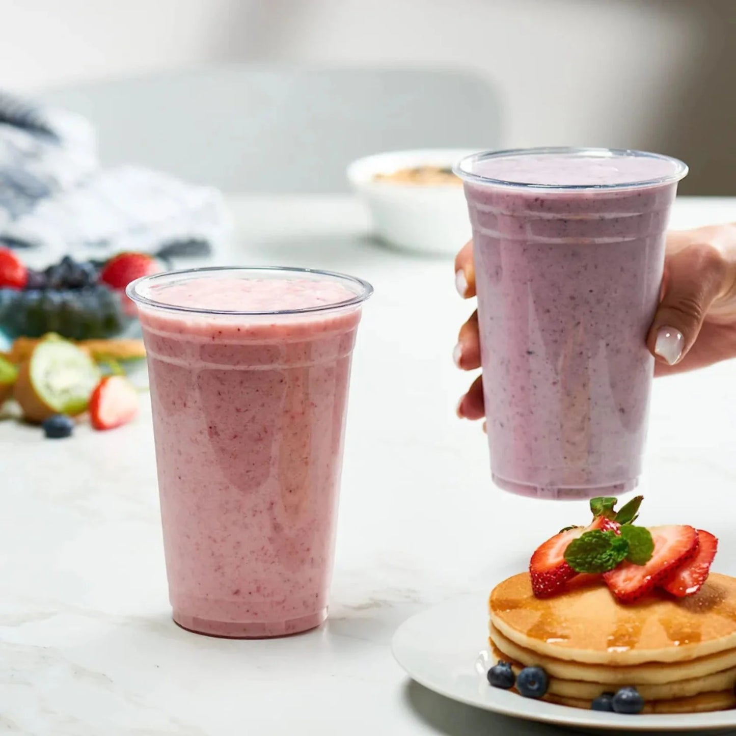 Two pink smoothies in plastic cups with a plate of pancakes topped with strawberries and blueberries on a white surface.