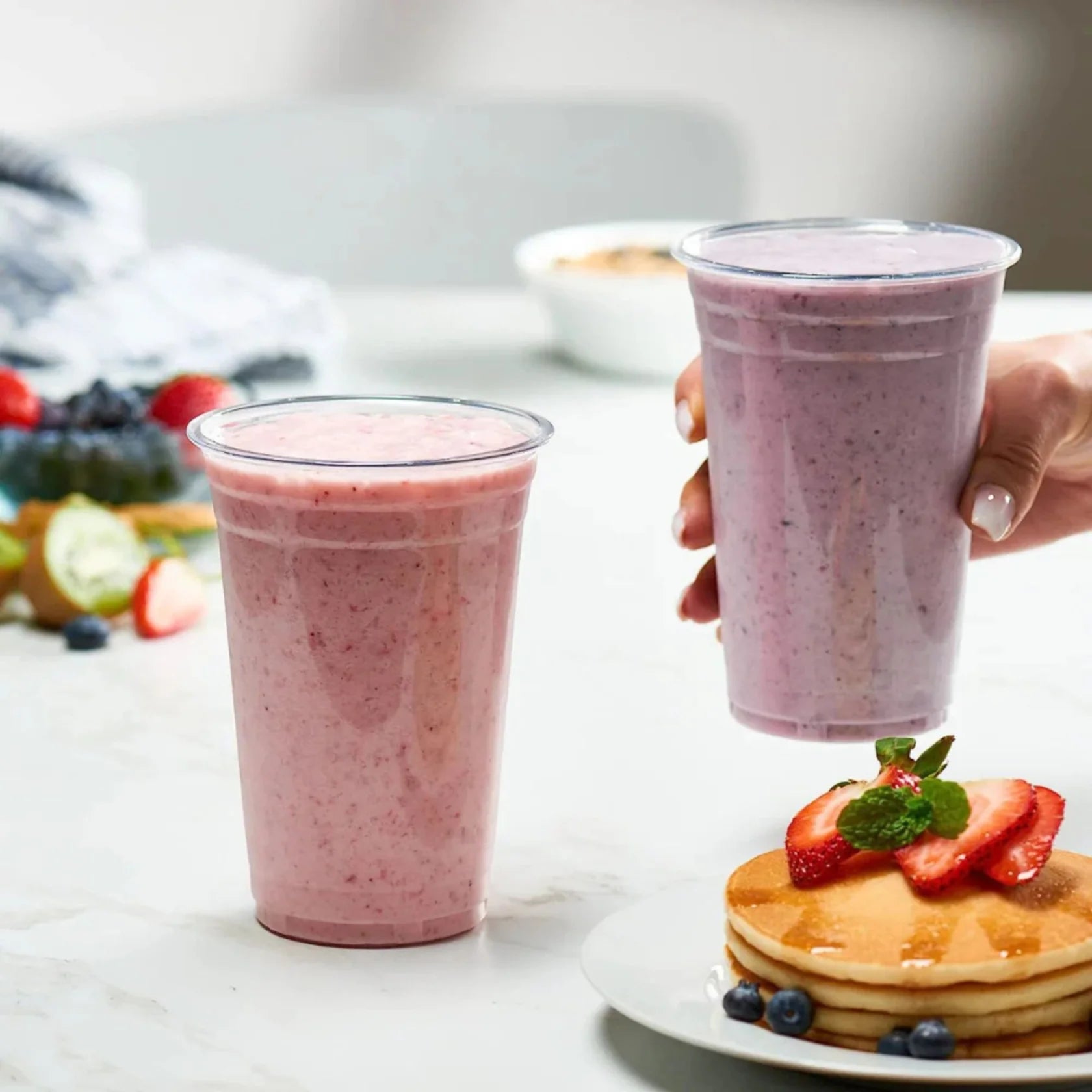 Two pink smoothies in plastic cups with a plate of pancakes topped with strawberries and blueberries on a white surface.