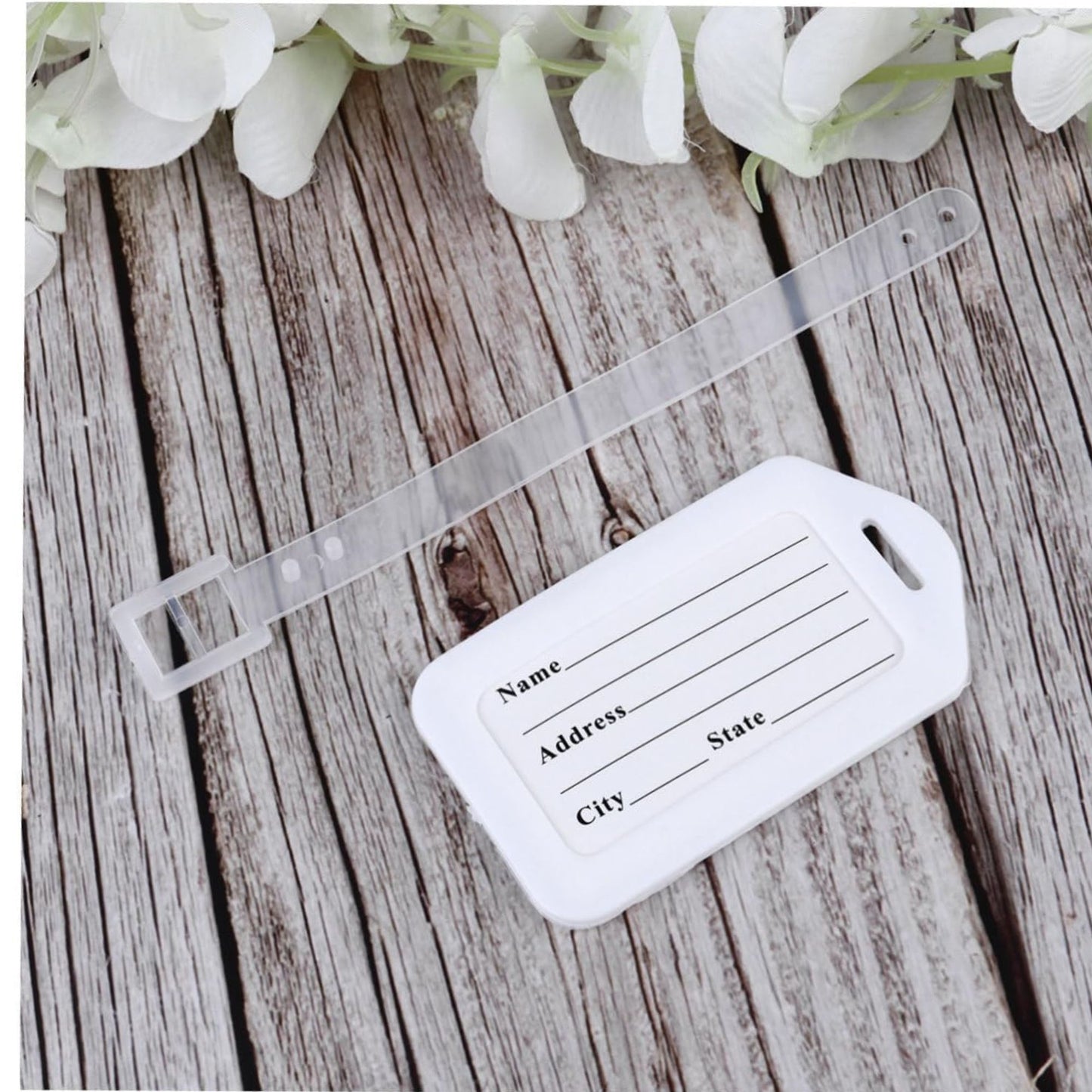 White luggage tag with text fields on a wooden surface with white flowers.