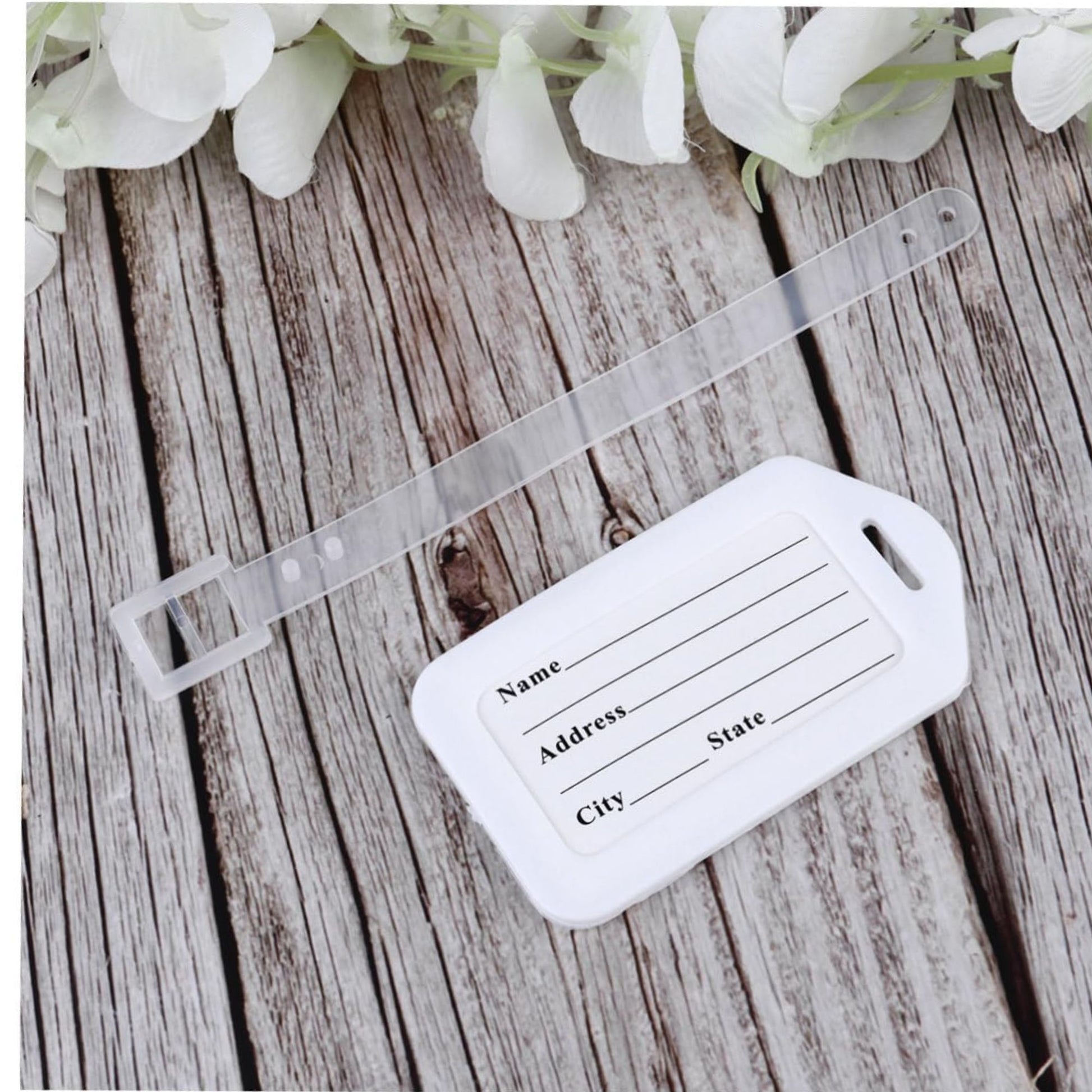 White luggage tag with text fields on a wooden surface with white flowers.