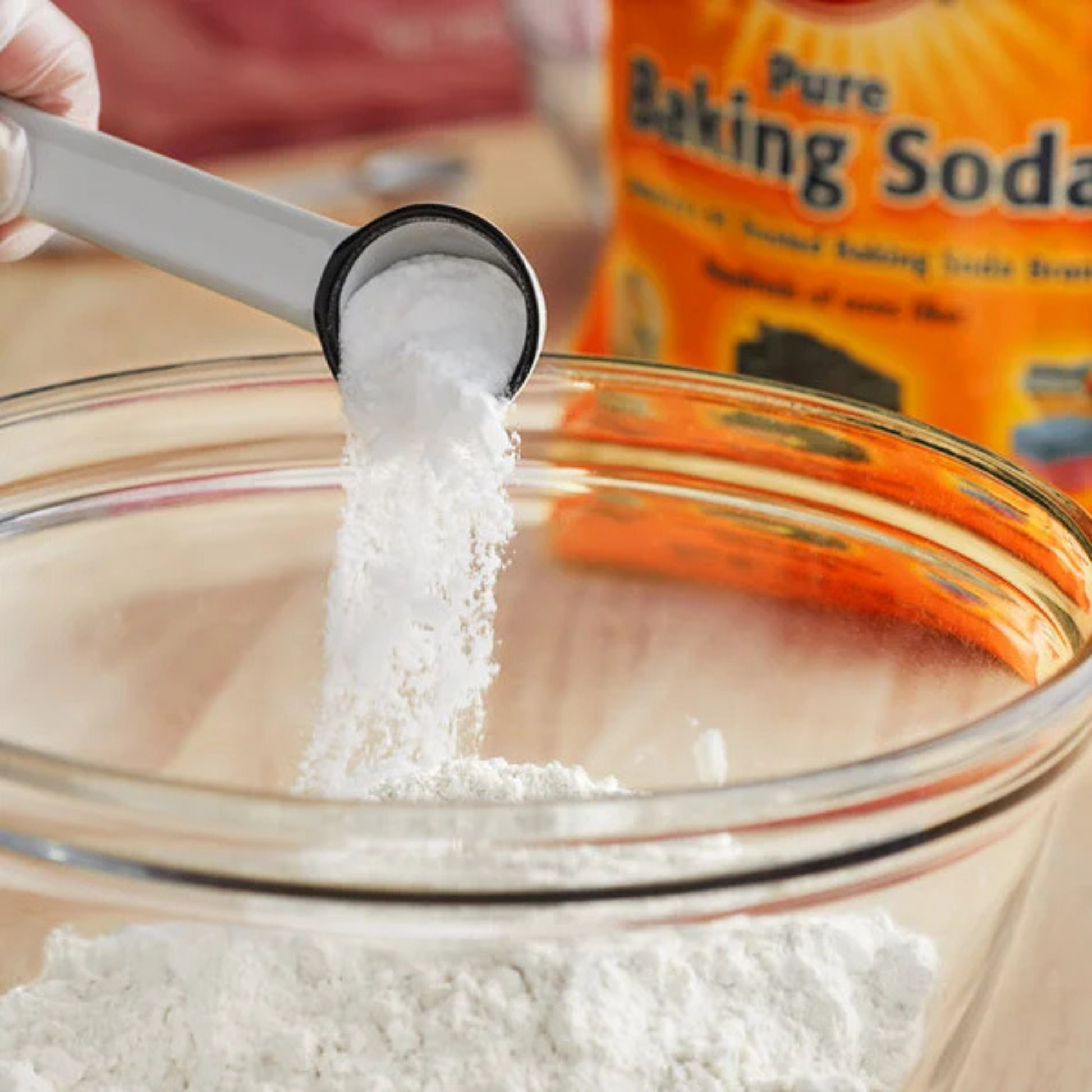 Baking soda being measured into a glass bowl with a container of baking soda in the background.